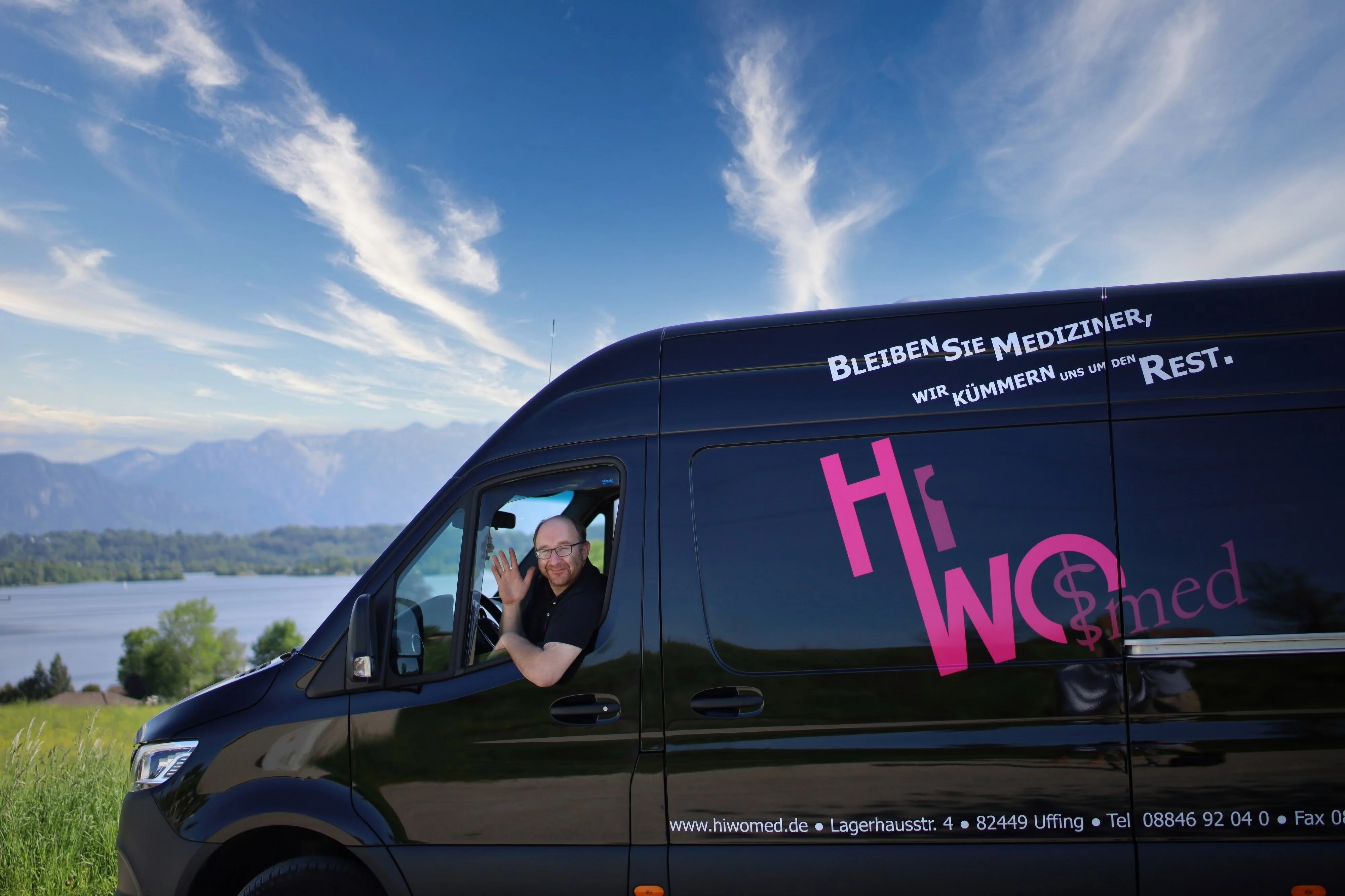 Ein HiWo-med-Mitarbeiter winkt aus dem Fenster seines Lieferfahrzeugs, im Hintergrund der Staffelsee mit Alpenkette.